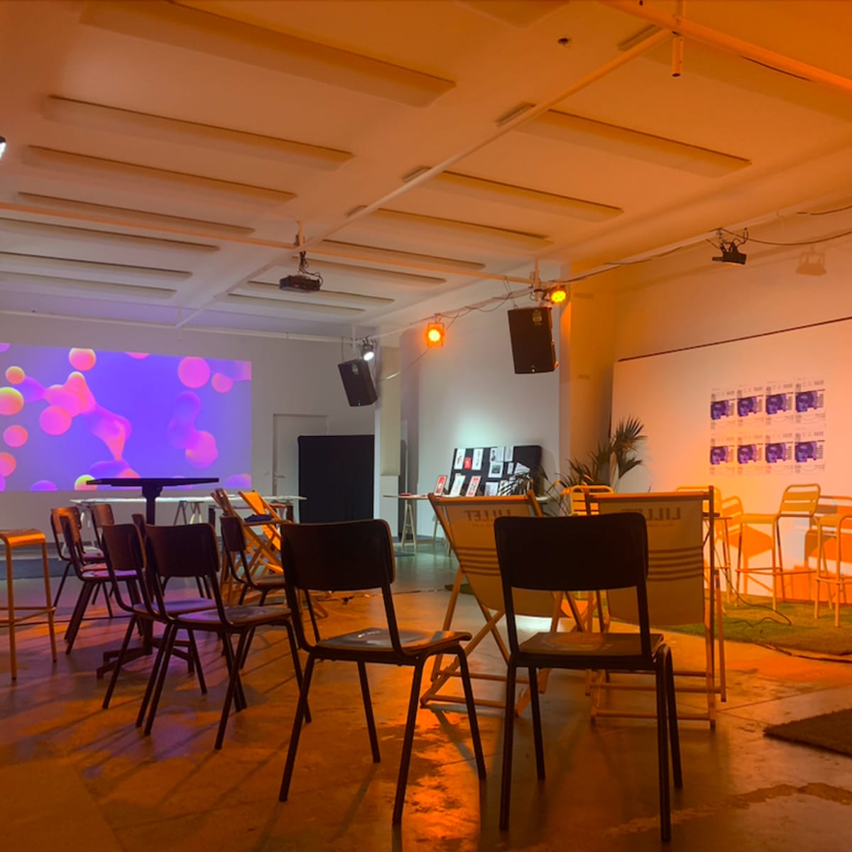 A spacious room with chairs arranged in rows facing a large screen displaying colorful visuals, complemented by a few potted plants and speakers mounted on the walls.