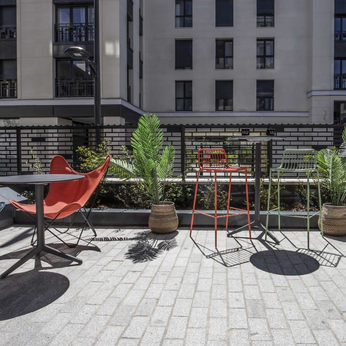 The scene depicts an outdoor terrace area with colorful furniture, including chairs and tables, surrounded by potted plants and a modern building backdrop.