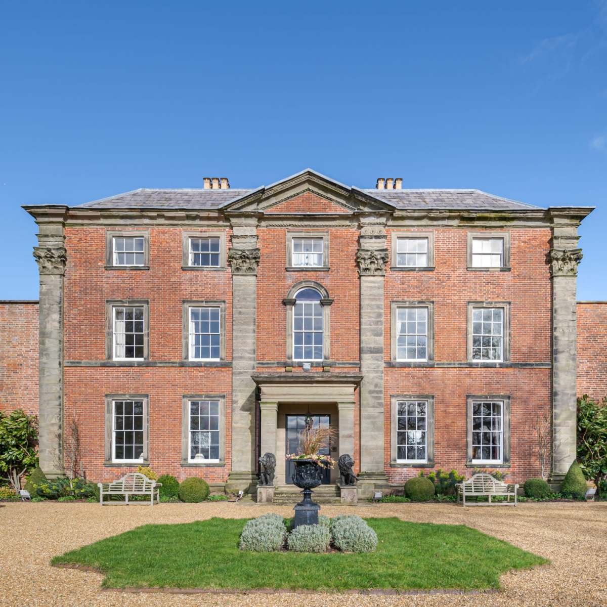 A large, red-brick mansion with symmetrical windows and a central entrance, surrounded by manicured gardens and gravel pathways.