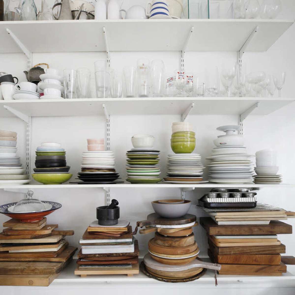 Shelves filled with various dishes, glassware, and kitchen accessories, along with a stack of wooden cutting boards on a surface below.