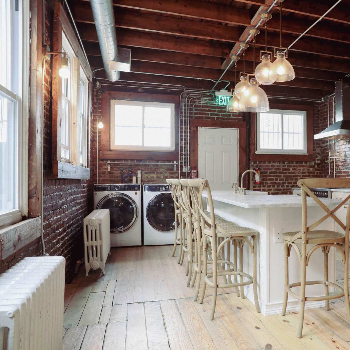 A modern kitchen with exposed brick walls, a large island with seating, and a washer and dryer located beside the refrigerator.