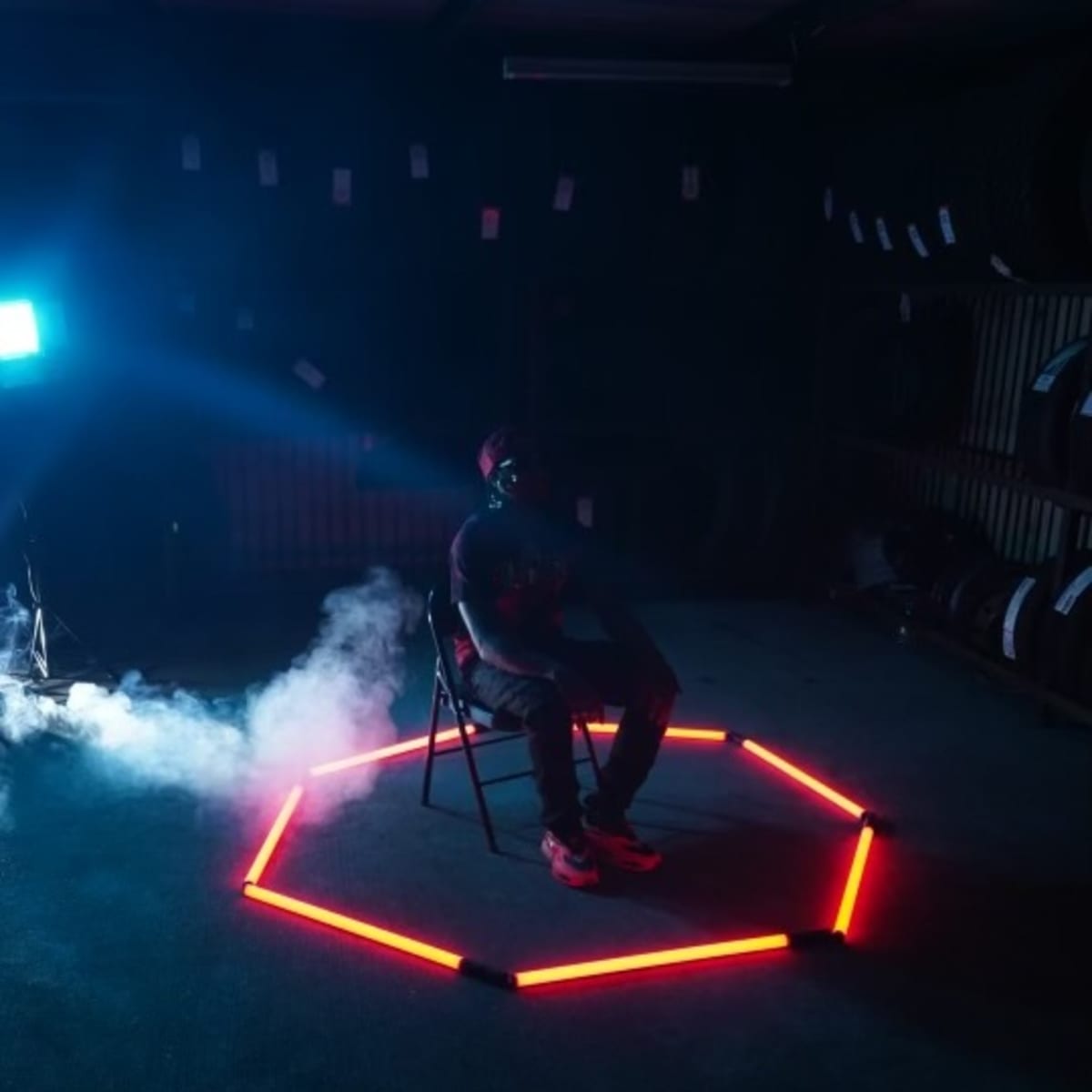 A person sits on a chair in the center of an illuminated hexagonal shape made of red lights, surrounded by smoke in a dimly-lit room.