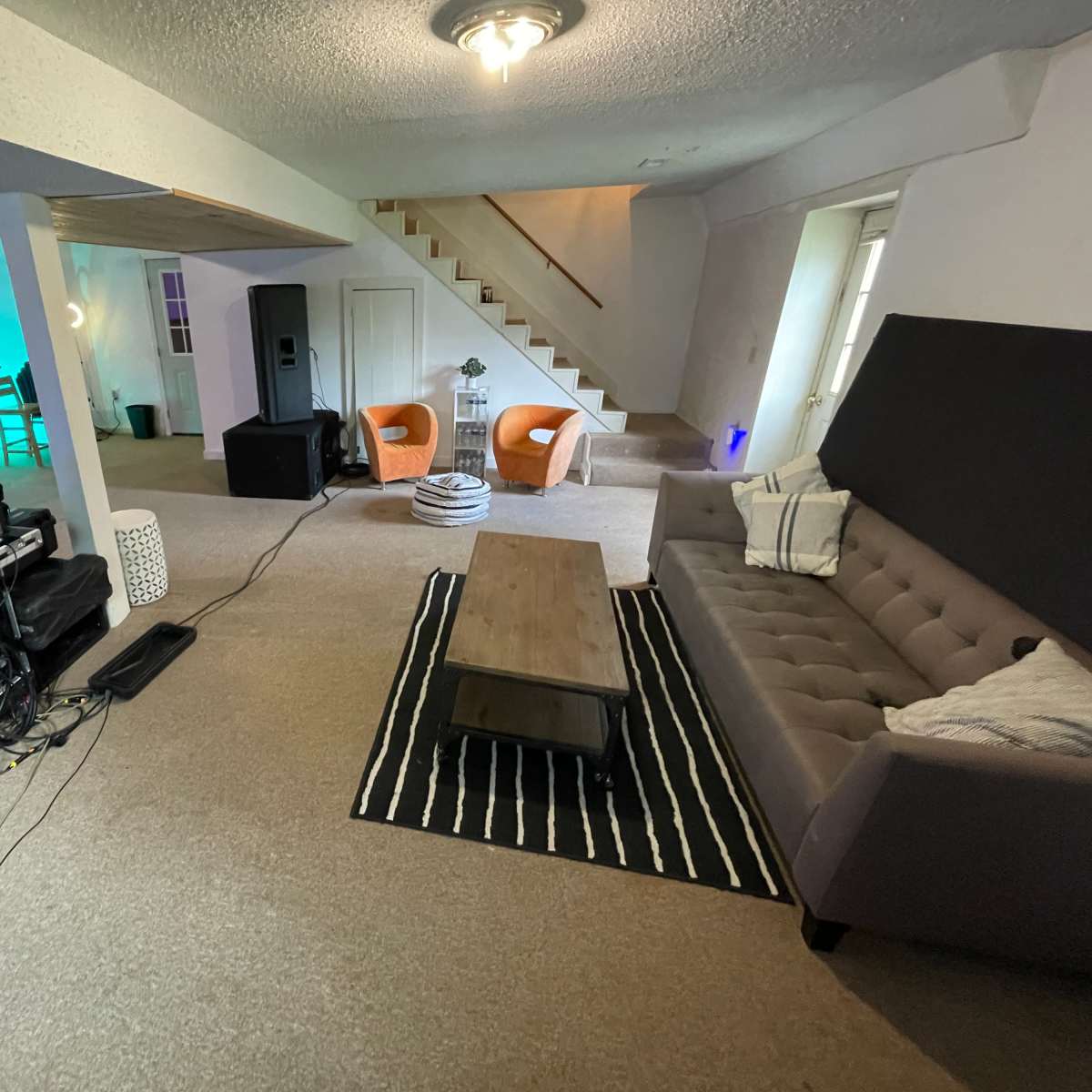 A spacious room with a recording setup on one side, a couch and chairs in the center, and a staircase leading to the upper floor.