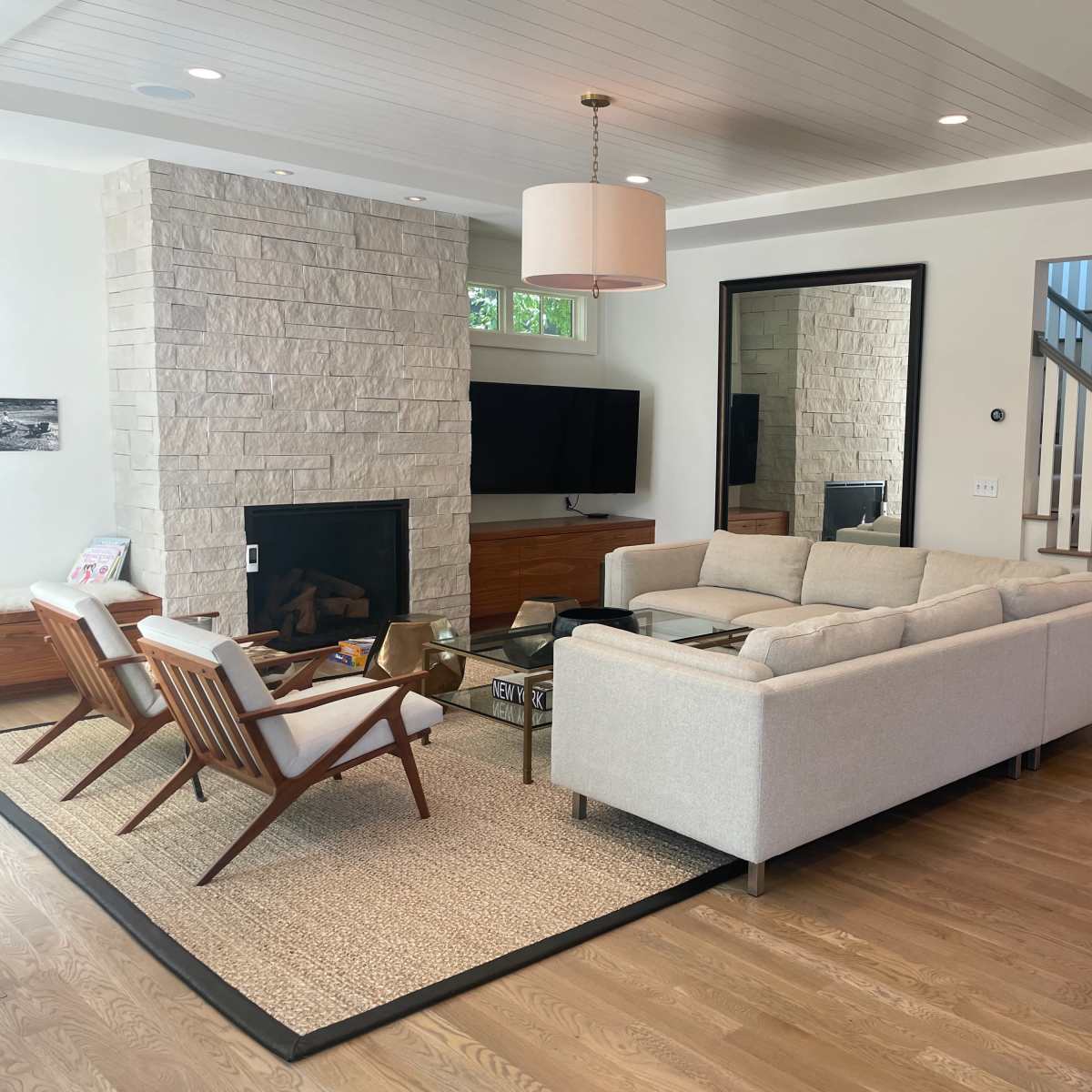 A modern living room with a light-colored sofa, a stone fireplace, a television mounted on the wall, and a staircase in the background.