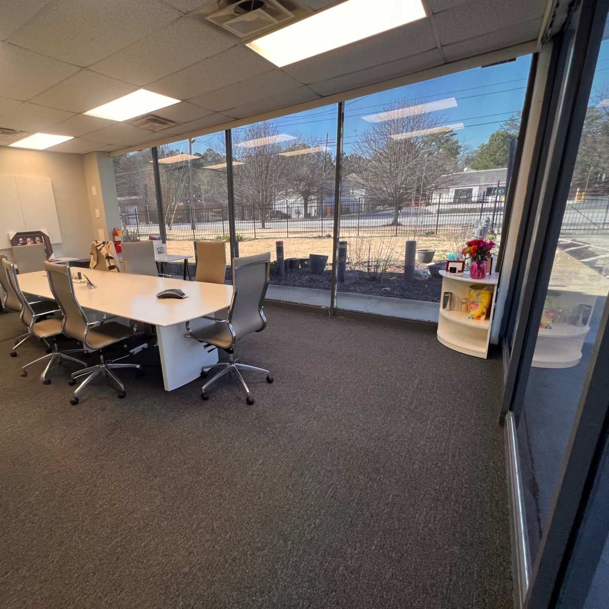 A conference room with a long table and several chairs, featuring large windows that provide a view of an outdoor area.