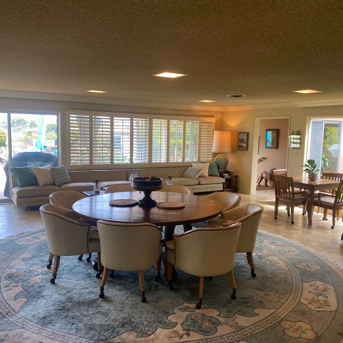 A spacious living area features a round dining table surrounded by chairs, with large windows providing natural light and views of the patio and garden.