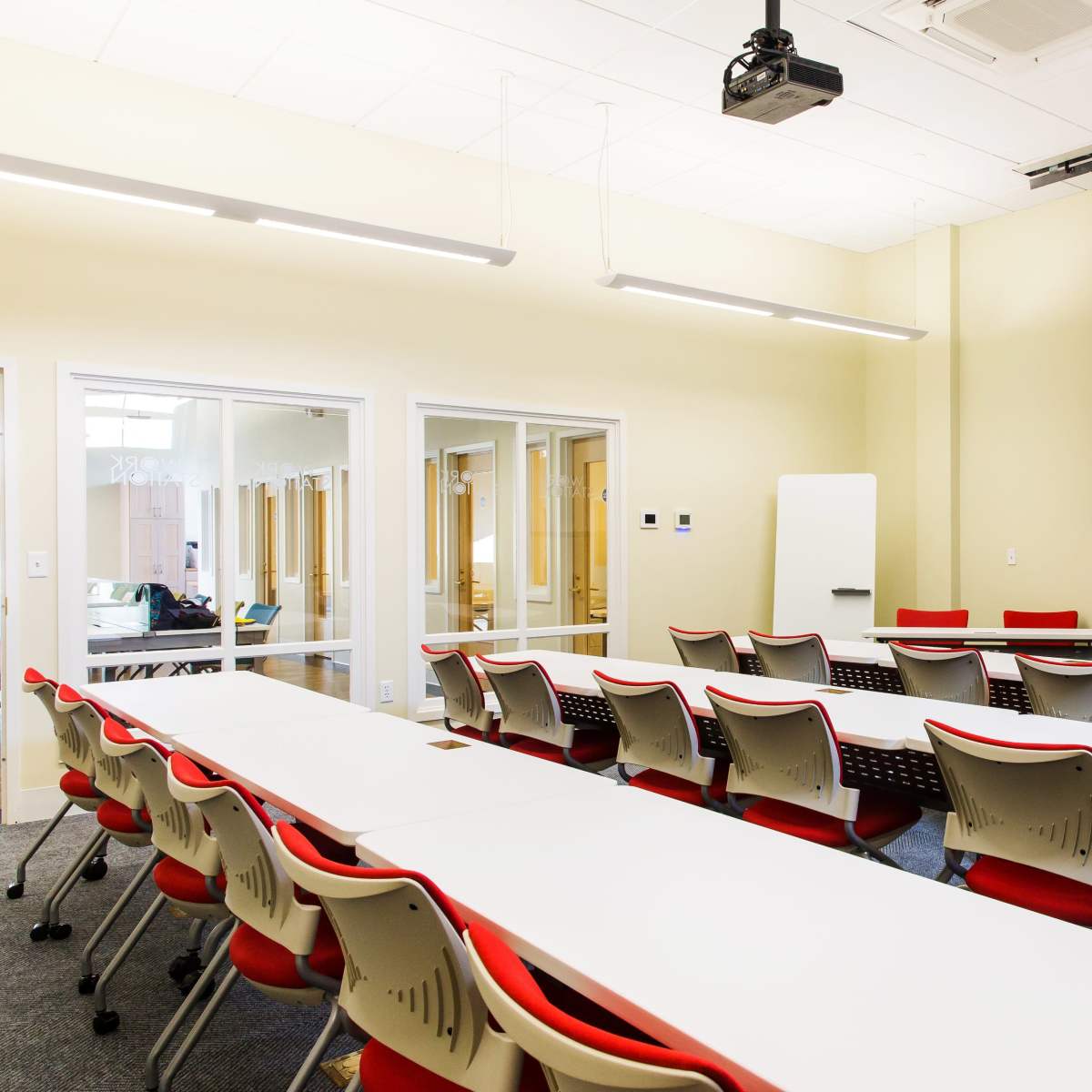 A classroom setting with rows of white tables and red chairs arranged facing a large projection screen on the wall.