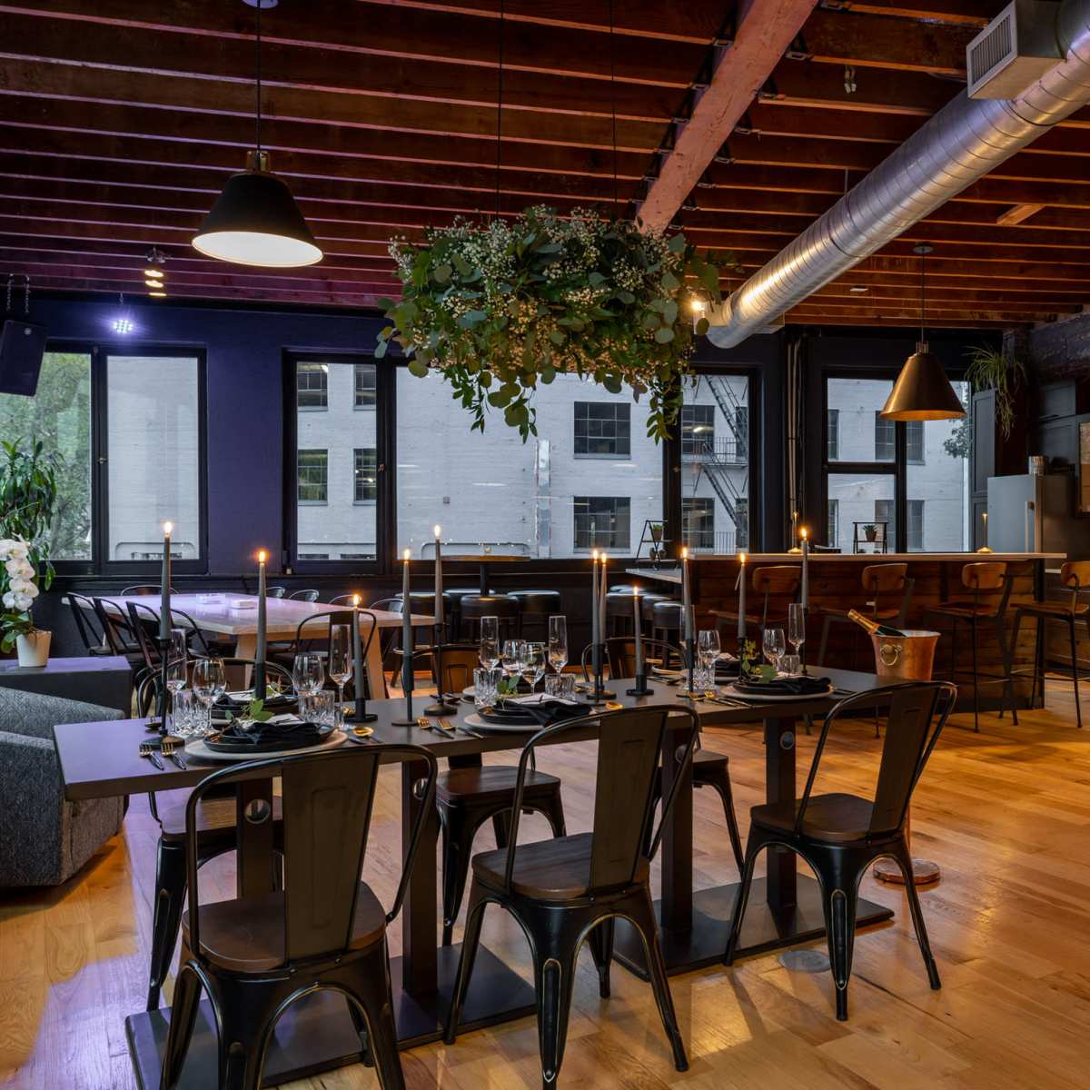 A modern dining space featuring wooden flooring, a large table set for dinner, and a bar area with sleek lighting and plants.
