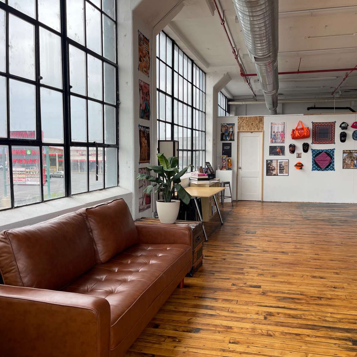A spacious room with large windows, featuring a brown leather couch, a desk, and a wall adorned with colorful artwork.