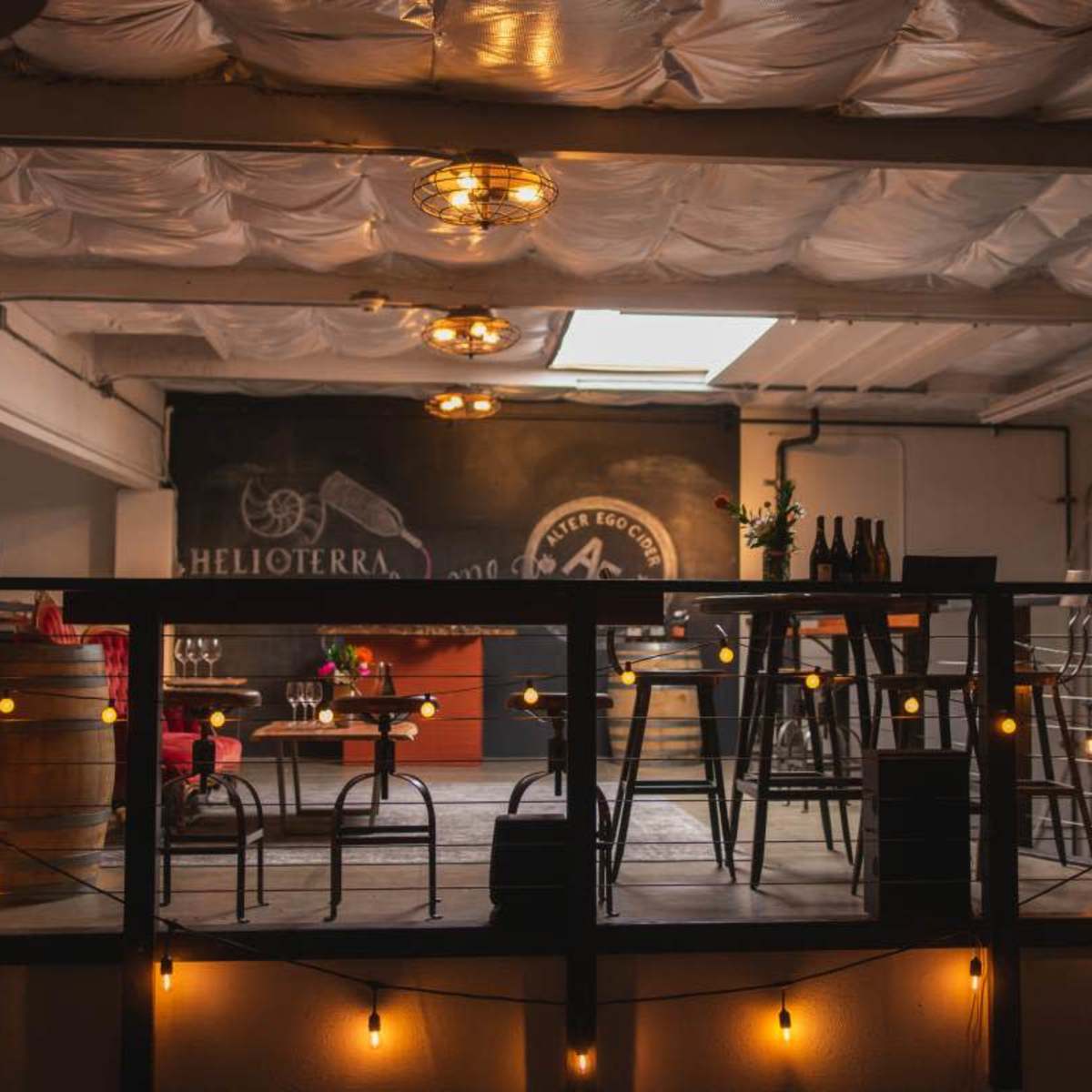 A dimly lit interior of a bar or event space, featuring a raised seating area with string lights and wooden furniture.