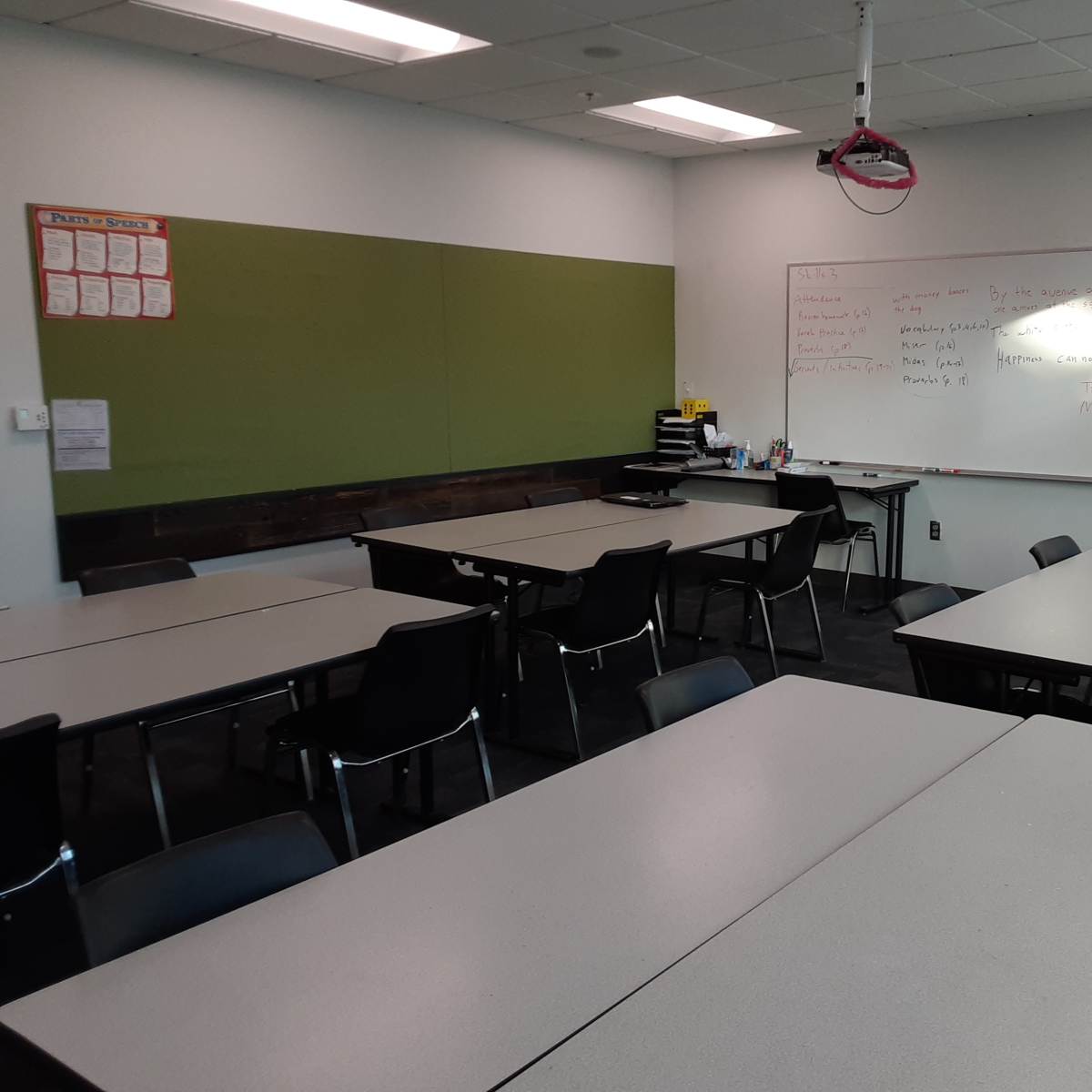 A classroom is set up with several tables and chairs arranged around a whiteboard and a green wall.