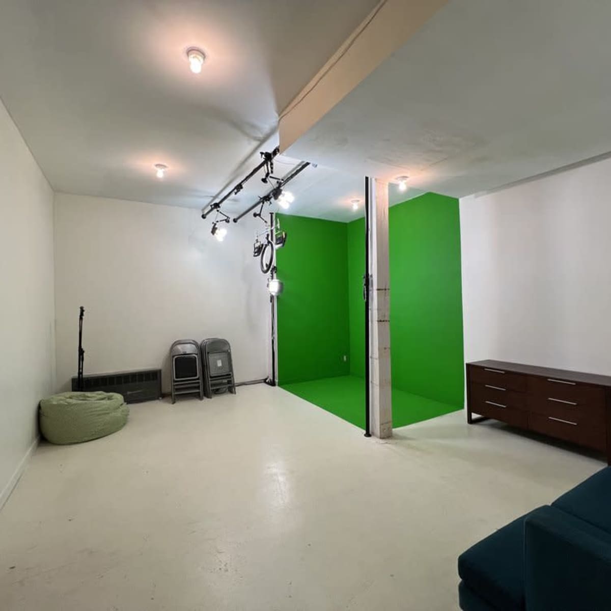 A spacious room featuring a green screen wall, a brown cabinet, two folding chairs, and a green bean bag on a white floor.