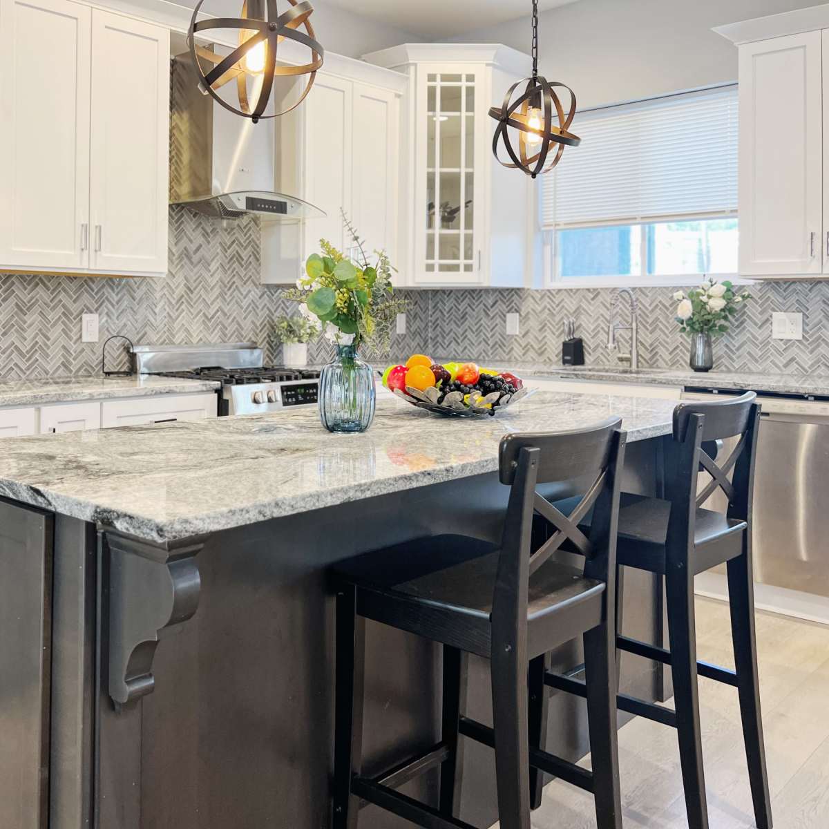 A modern kitchen features white cabinets, a gray marble countertop, and a central island with two black bar stools, surrounded by decorative items and fresh fruit.