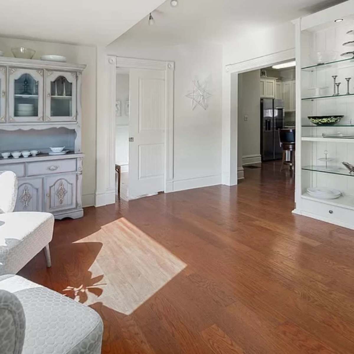 A brightly lit living space featuring a light-colored cabinet and a gray armchair, with wooden flooring and a doorway leading to a kitchen area.