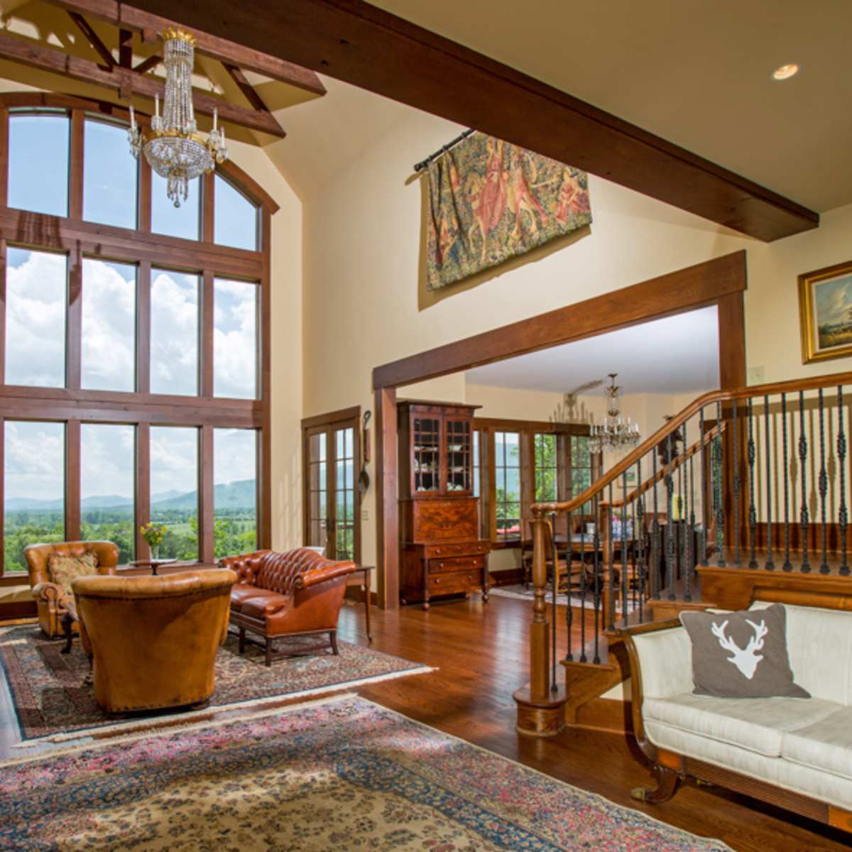 The spacious living room features large windows that frame a mountainous view, with a chandelier hanging above leather seating and a stone fireplace.