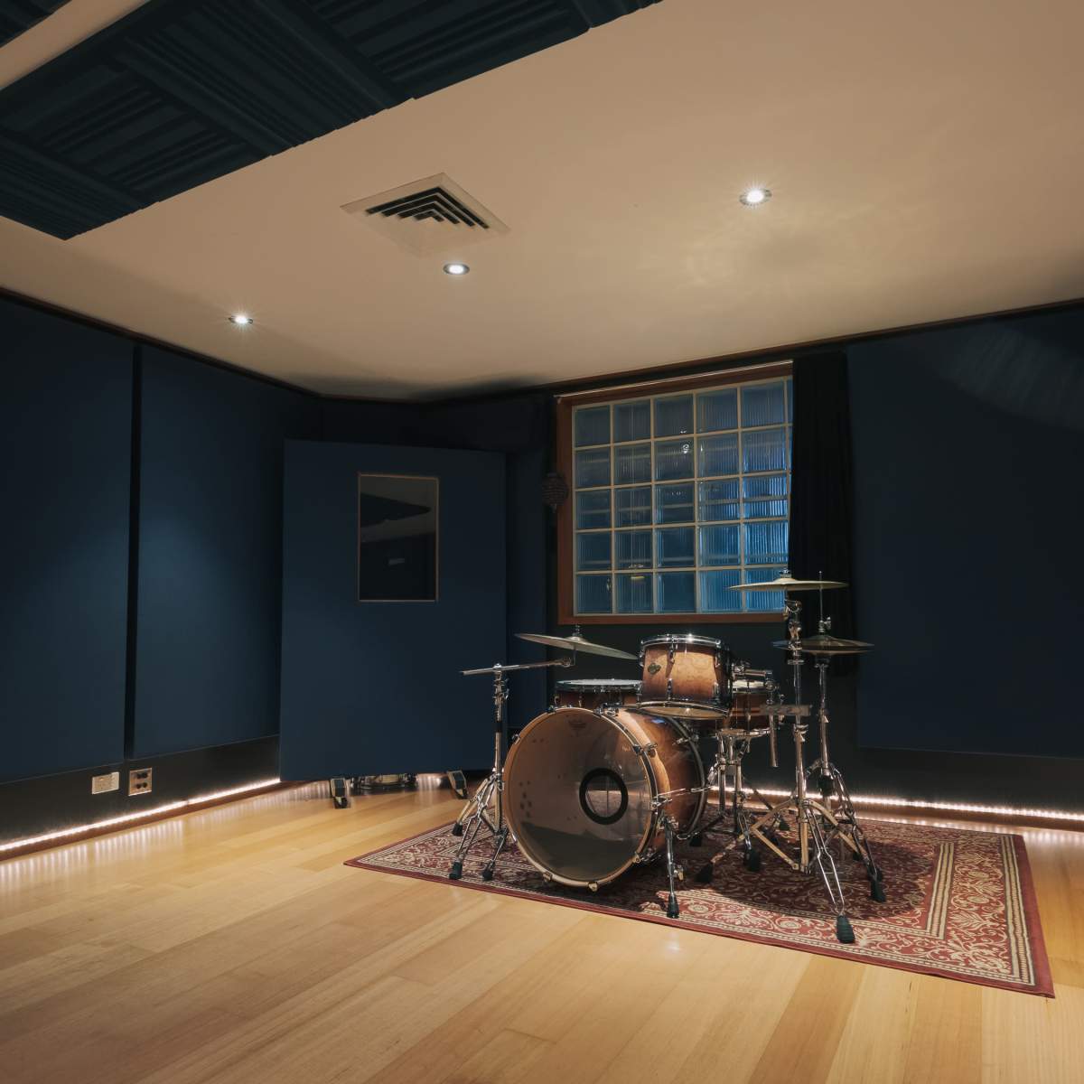 A music recording studio featuring a drum set positioned on a patterned rug in a room with dark blue walls and hardwood flooring.