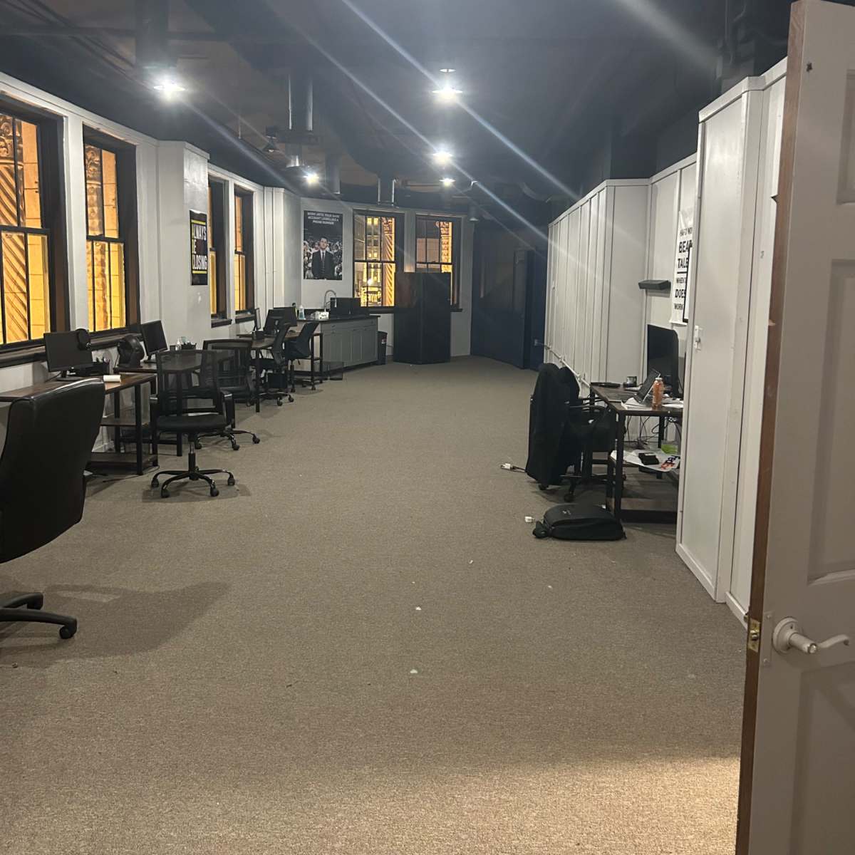 A dimly lit office corridor with several chairs and desks on either side, leading to a darker area at the far end.
