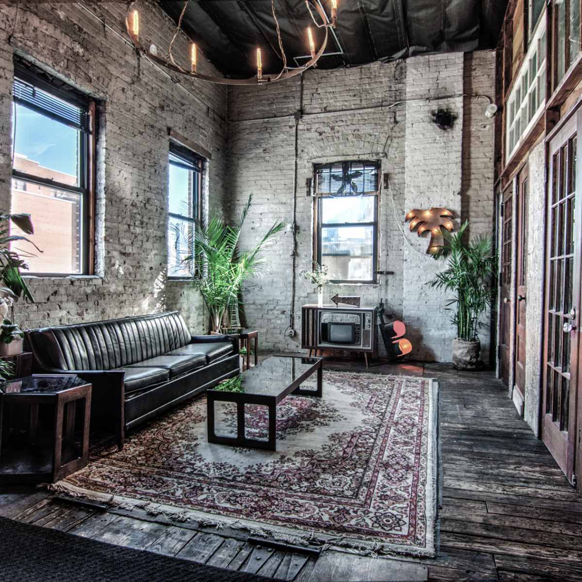 A spacious room with exposed brick walls, a leather sofa, a coffee table on a patterned rug, and large windows allowing natural light to enter.