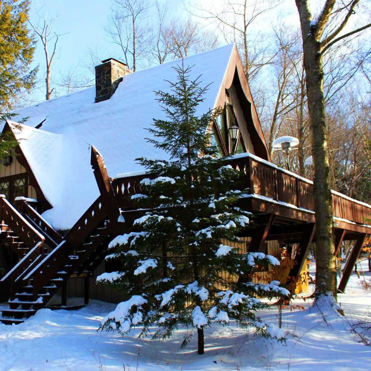 A large, A-frame cabin with snowy roofing and a wooden deck is surrounded by trees in a winter landscape.