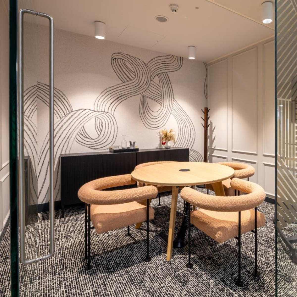 A small meeting room features a round table surrounded by four cushioned chairs, with a patterned wall design and a textured floor.