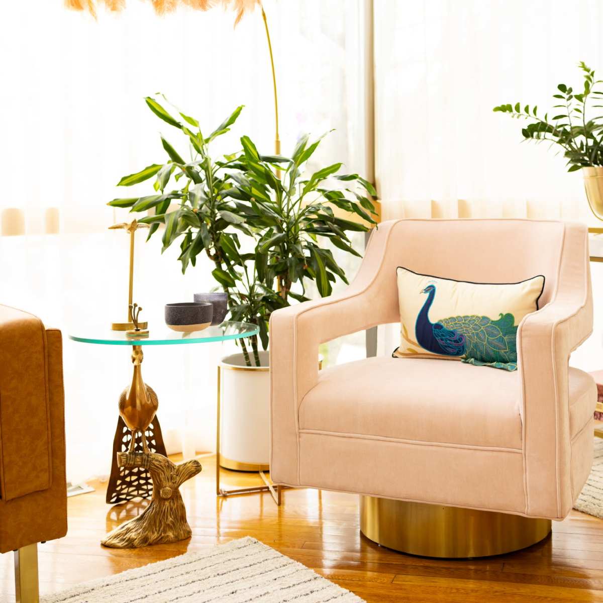 A soft pink armchair is positioned beside a glass side table adorned with plants and a decorative pillow featuring a peacock design.