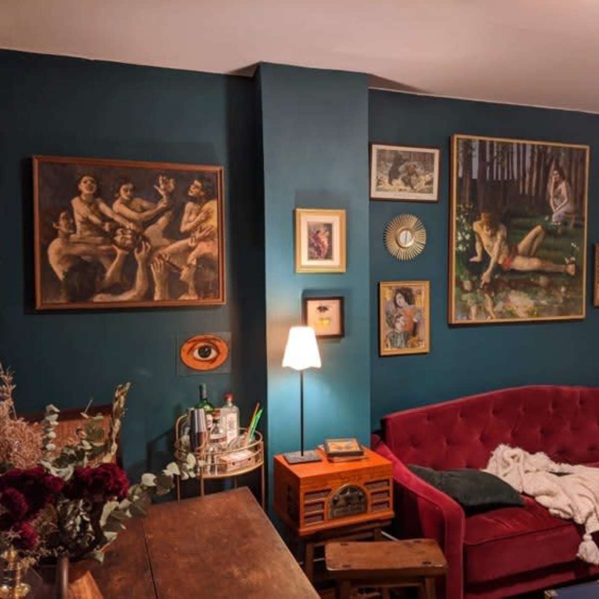 A living room featuring a red velvet couch, a wooden table, and a wall adorned with various framed paintings against a teal backdrop.