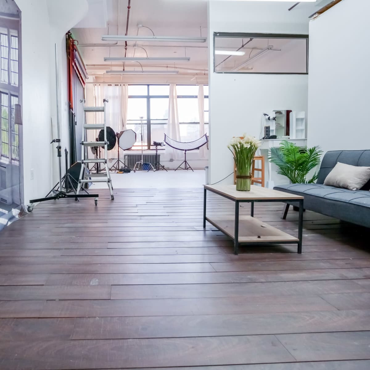 A spacious, well-lit studio featuring wooden flooring, large windows, and photography equipment arranged in the background.