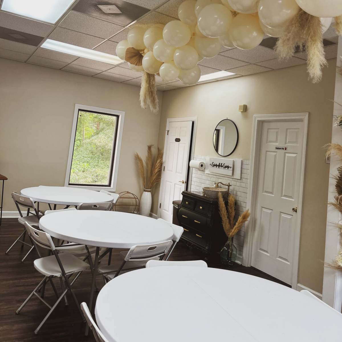 A series of beige balloons is arranged overhead in a room with white tables and chairs, featuring a window and a decorative wall.