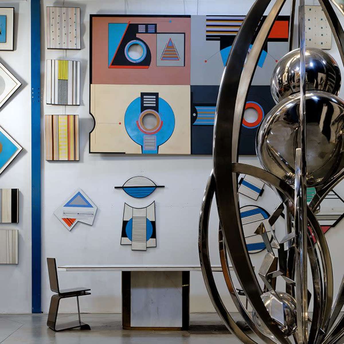 A contemporary art gallery with abstract wall paintings and a metallic sculpture in the foreground near a minimalist table and chair.