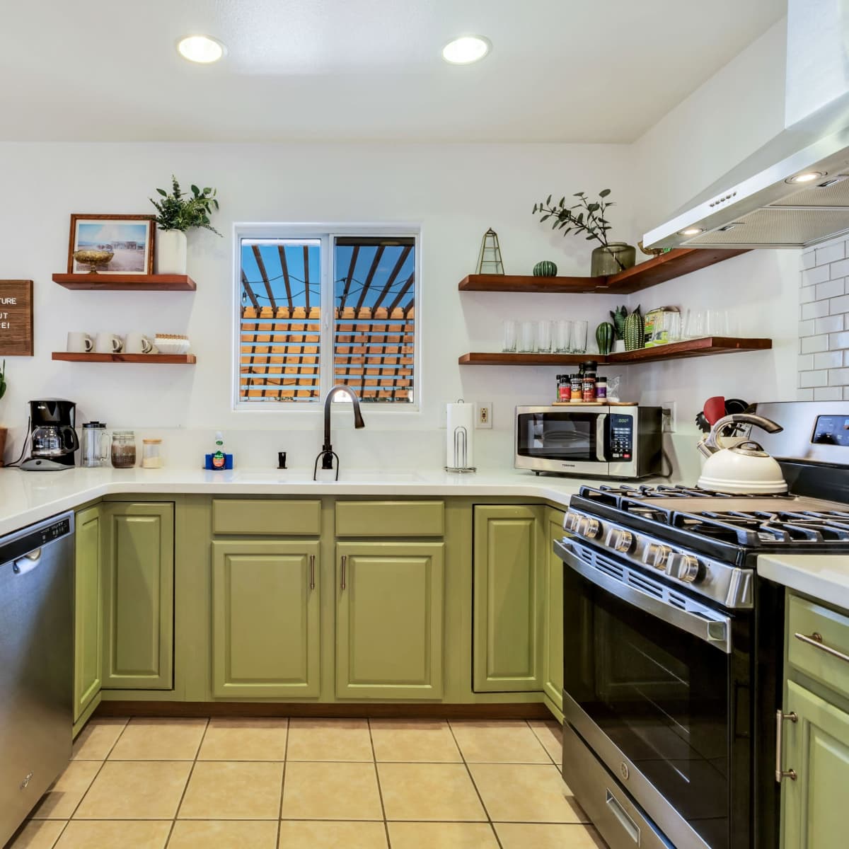 The kitchen features green cabinets, a white countertop, and stainless steel appliances with open shelving displaying plants and kitchenware.