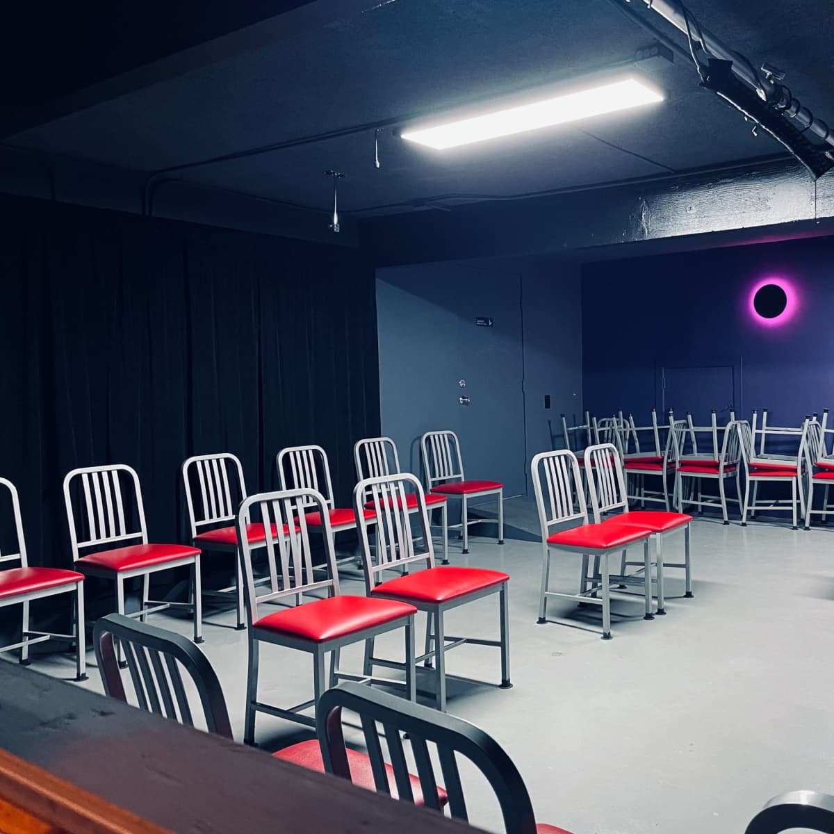A dimly lit room with a set of metal chairs arranged around a small stage, featuring a microphone stand and a purple wall with circular lighting.