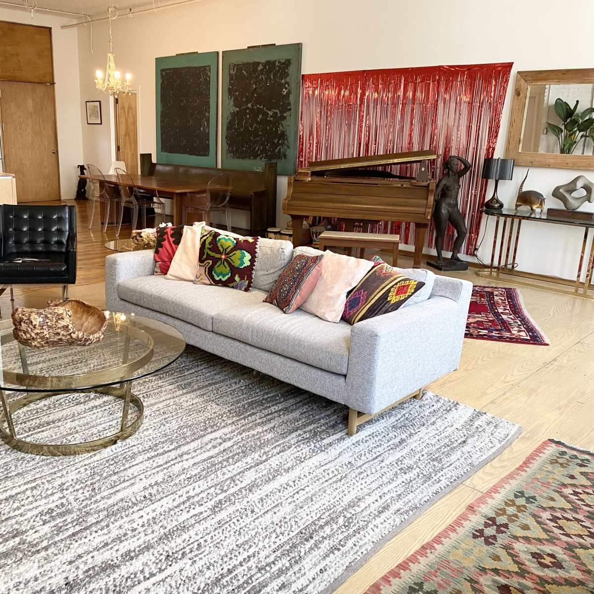 A modern living space featuring a gray sofa with colorful cushions, a round glass coffee table, and a piano, surrounded by a mix of decorative rugs and artwork.