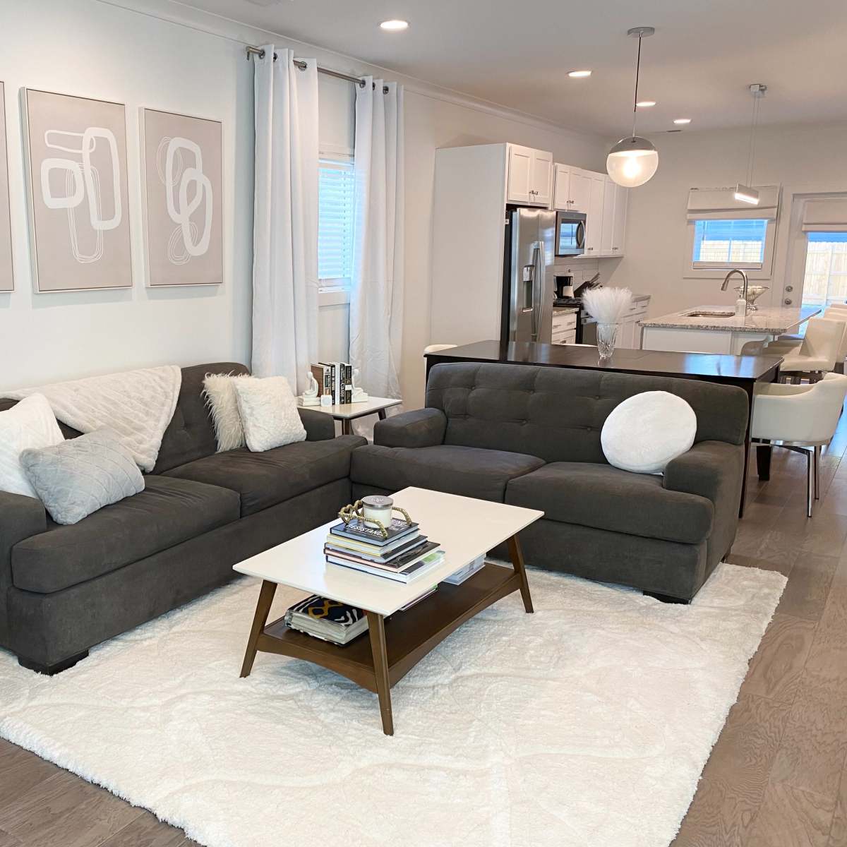A modern living room featuring a dark sectional sofa, a white coffee table, and framed abstract artwork on the wall, with a kitchen visible in the background.