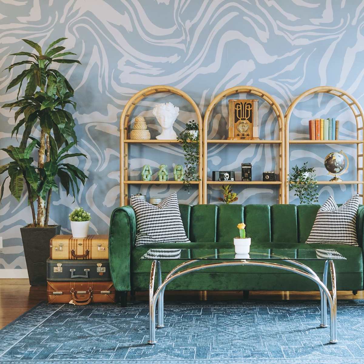 A green velvet sofa is positioned in front of a light blue marbled wall, accompanied by a glass coffee table and decorated shelves filled with various items, plants, and a patterned area rug.