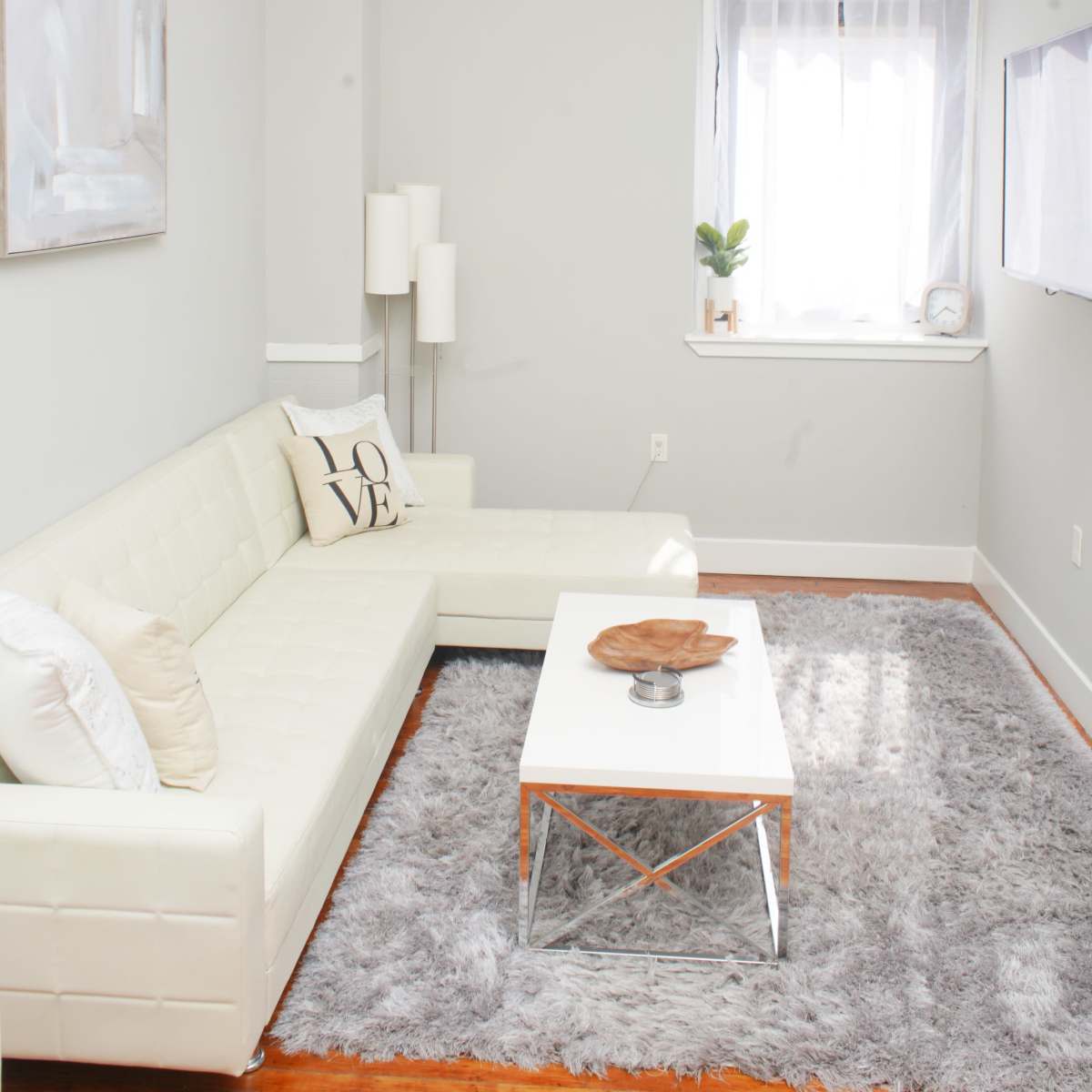 A modern living room features a white leather sectional sofa, a glass coffee table, and a large area rug, with a window allowing natural light to enter.