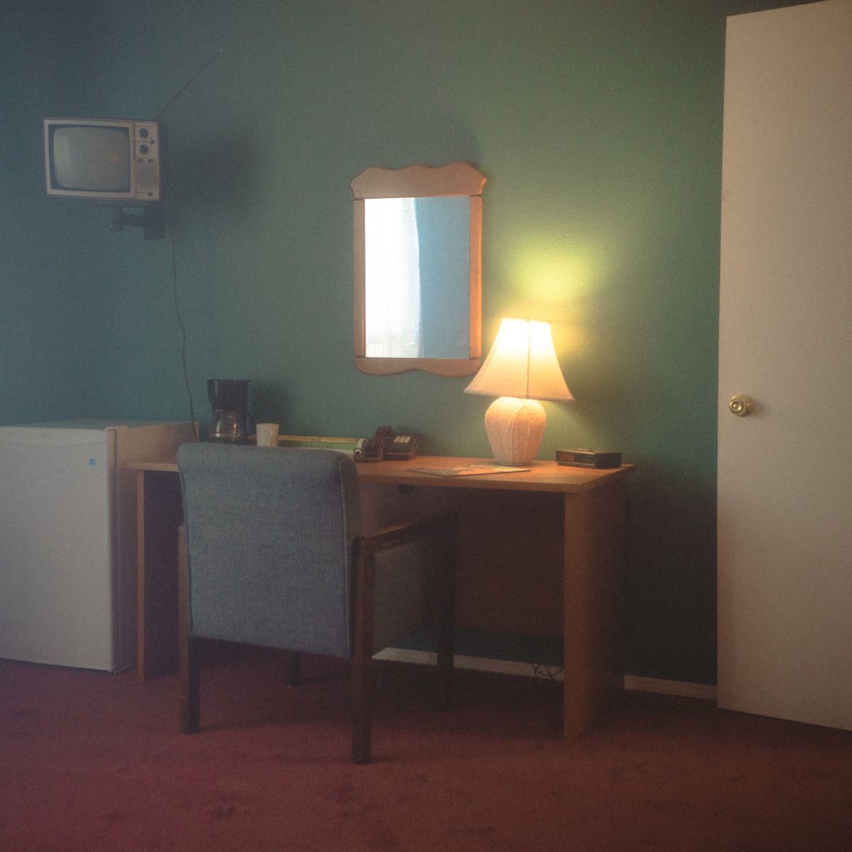 A small, simple hotel room with a green wall, a desk with a chair, a lamp, a mirror, a television, a refrigerator, and a door numbered four.