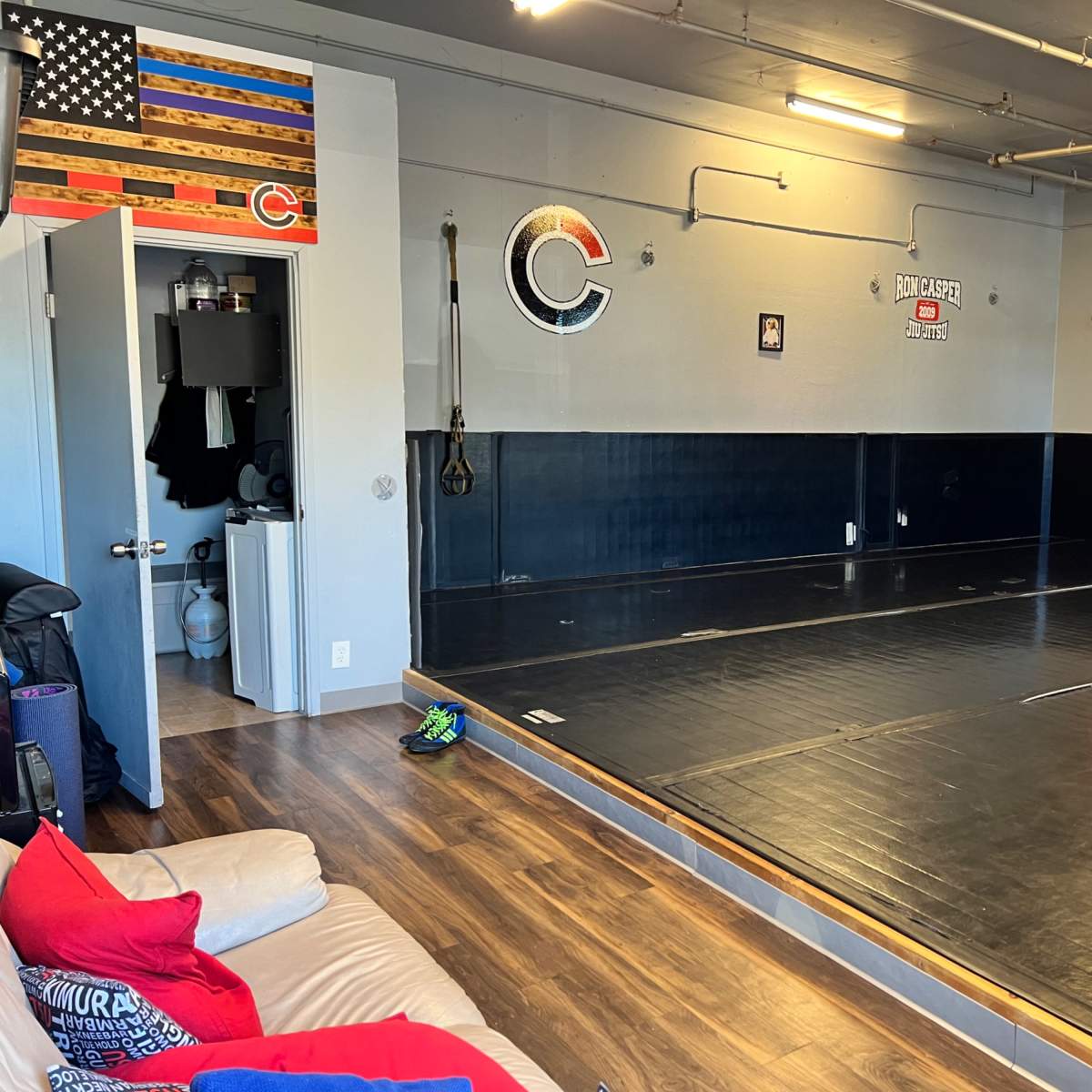 A training room with a large, black mat space, an American flag on the wall, and a doorway leading to a restroom area.