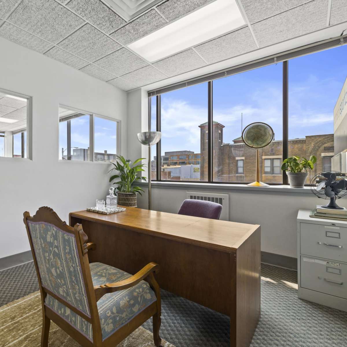 A bright office space featuring a wooden desk, an armchair, and a large window with a view of a cityscape.