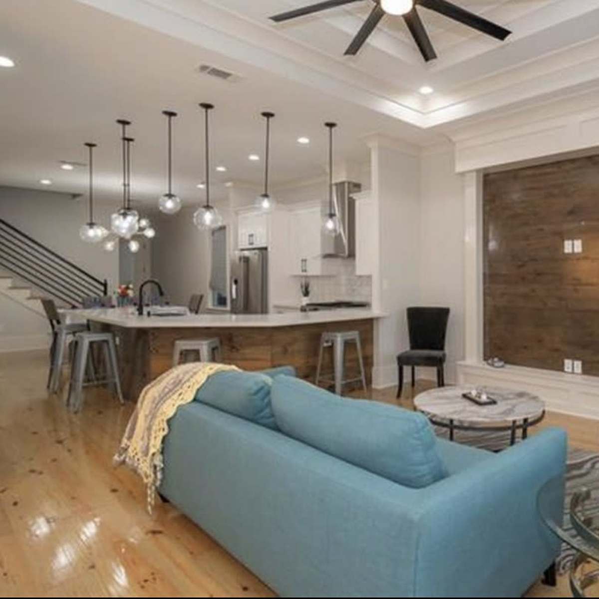 A modern living room with a light-colored sofa, a dark blue armchair, a round coffee table, and a kitchen area featuring pendant lights and stainless steel appliances.