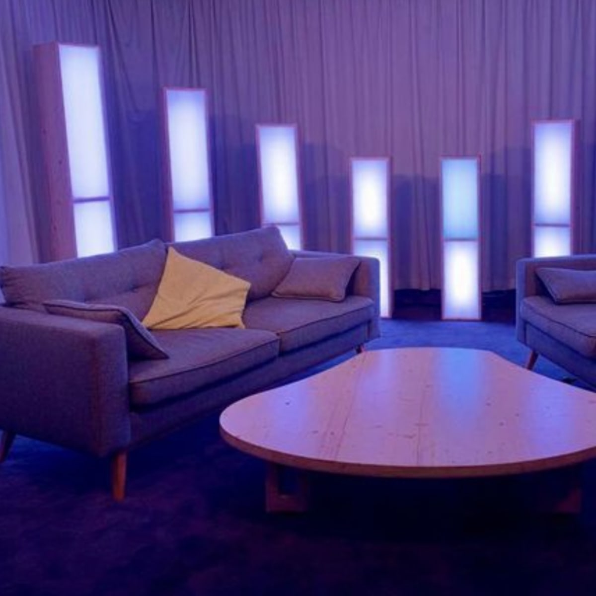Two gray sofas facing each other across a wooden coffee table, surrounded by illuminated vertical panels in a softly lit room.