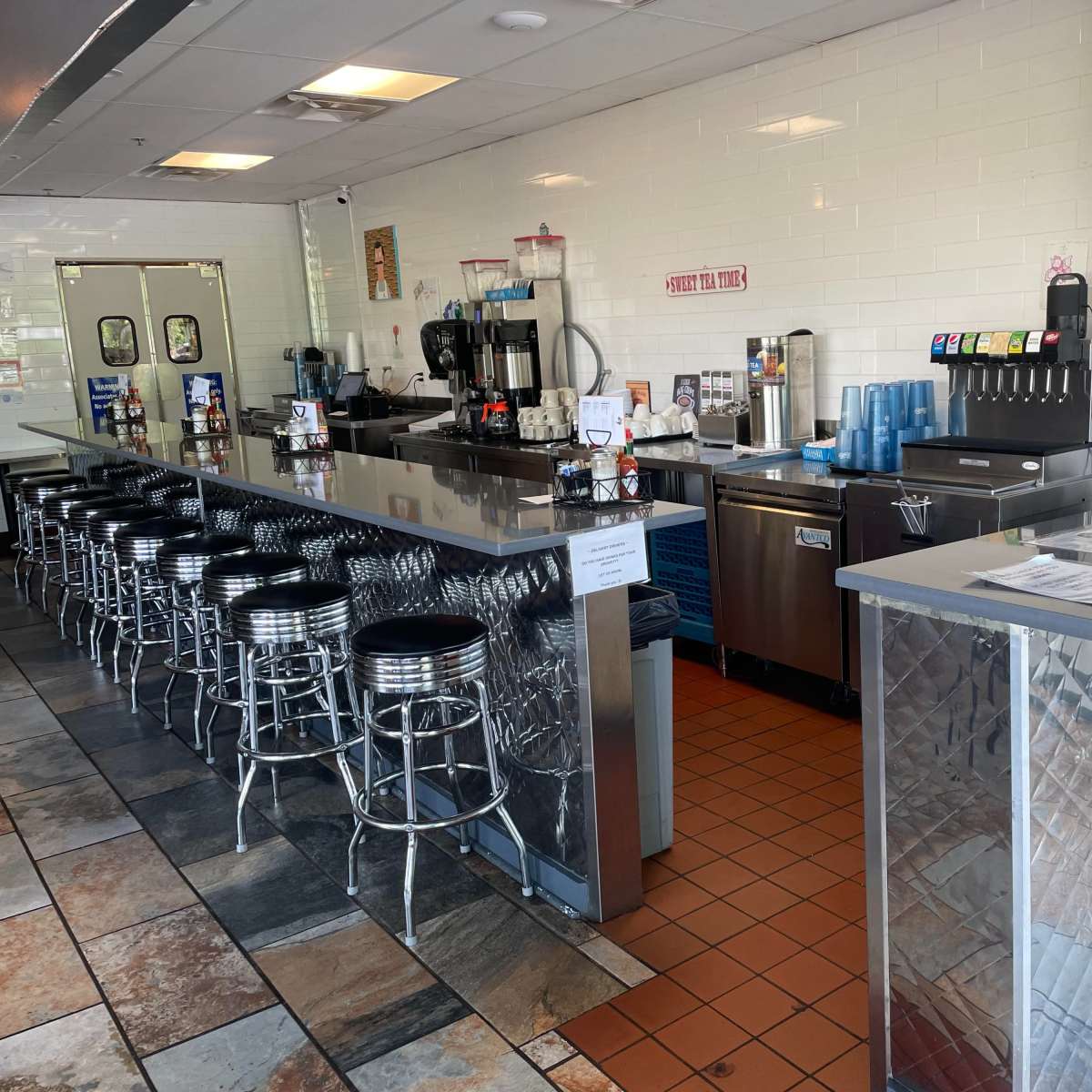 A diner-style interior with a long, silver bar counter lined with black stools, a drink dispenser, and various food preparation equipment in a tiled space.