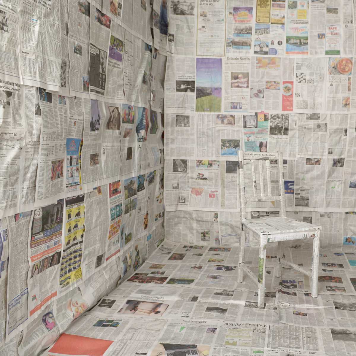 A single white chair is placed in the center of a room where the walls and floor are covered entirely in newspaper.