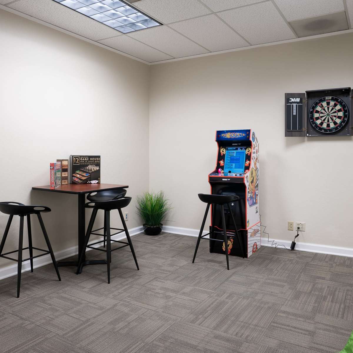 A small recreational room with a table and stools, a dartboard on the wall, an arcade machine, and green bean bags on the floor.