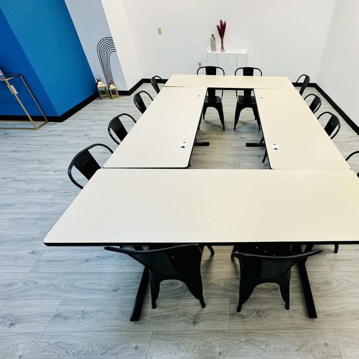 A rectangular arrangement of tables surrounded by black chairs is set up in a room with light-colored flooring and minimal decor.