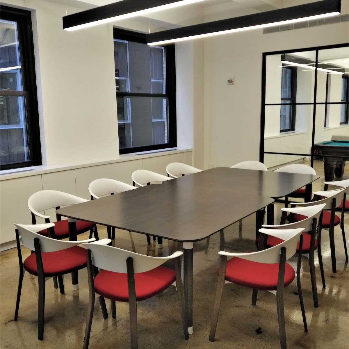 A modern conference room features a rectangular wooden table surrounded by eight chairs with white backs and red cushions, set against a backdrop of large windows and a mirror.