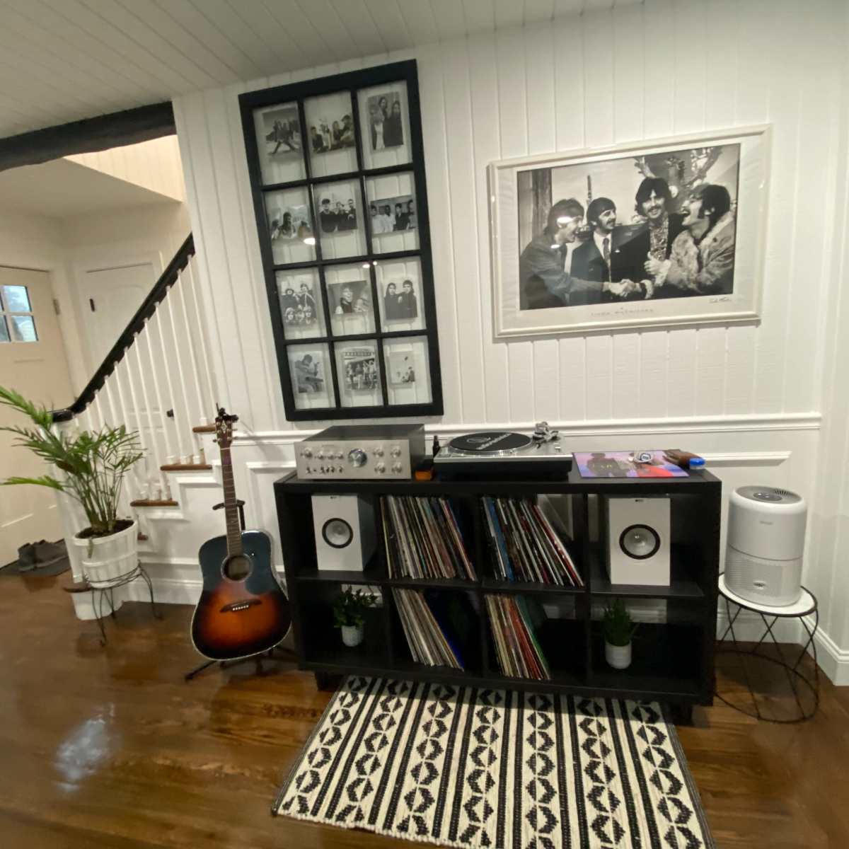 A living space with a black shelving unit holding vinyl records, an acoustic guitar, and a turntable, adjacent to a staircase and framed black-and-white photos on the wall.