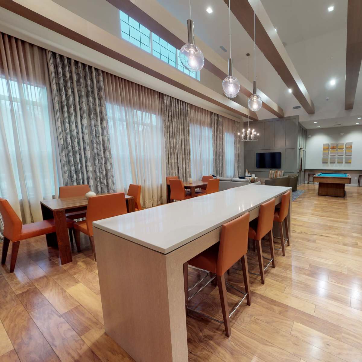 A modern dining area with a long central island, surrounded by orange chairs, leading to a spacious living area with wooden flooring and large windows.