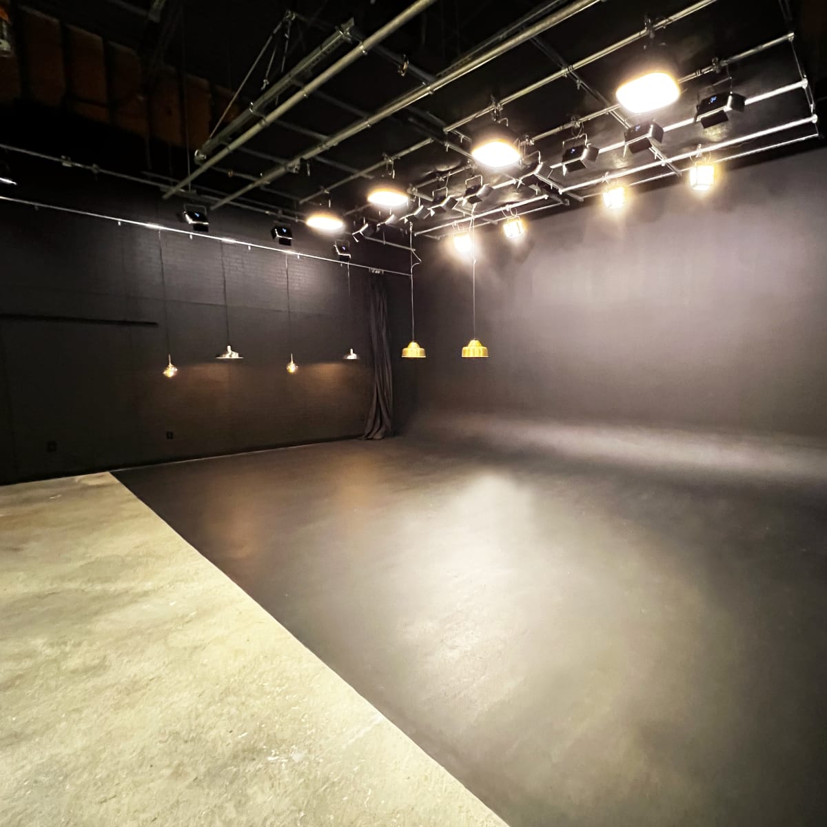 A large empty studio space with a black floor and walls, illuminated by overhead lights.