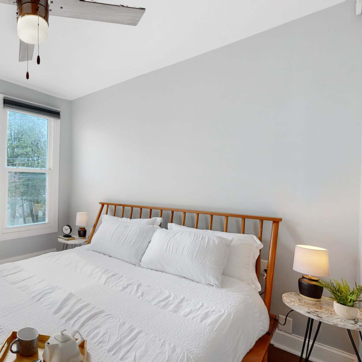 A well-lit bedroom features a queen-sized bed with white bedding, a wooden headboard, two windows, and framed artwork on the wall.