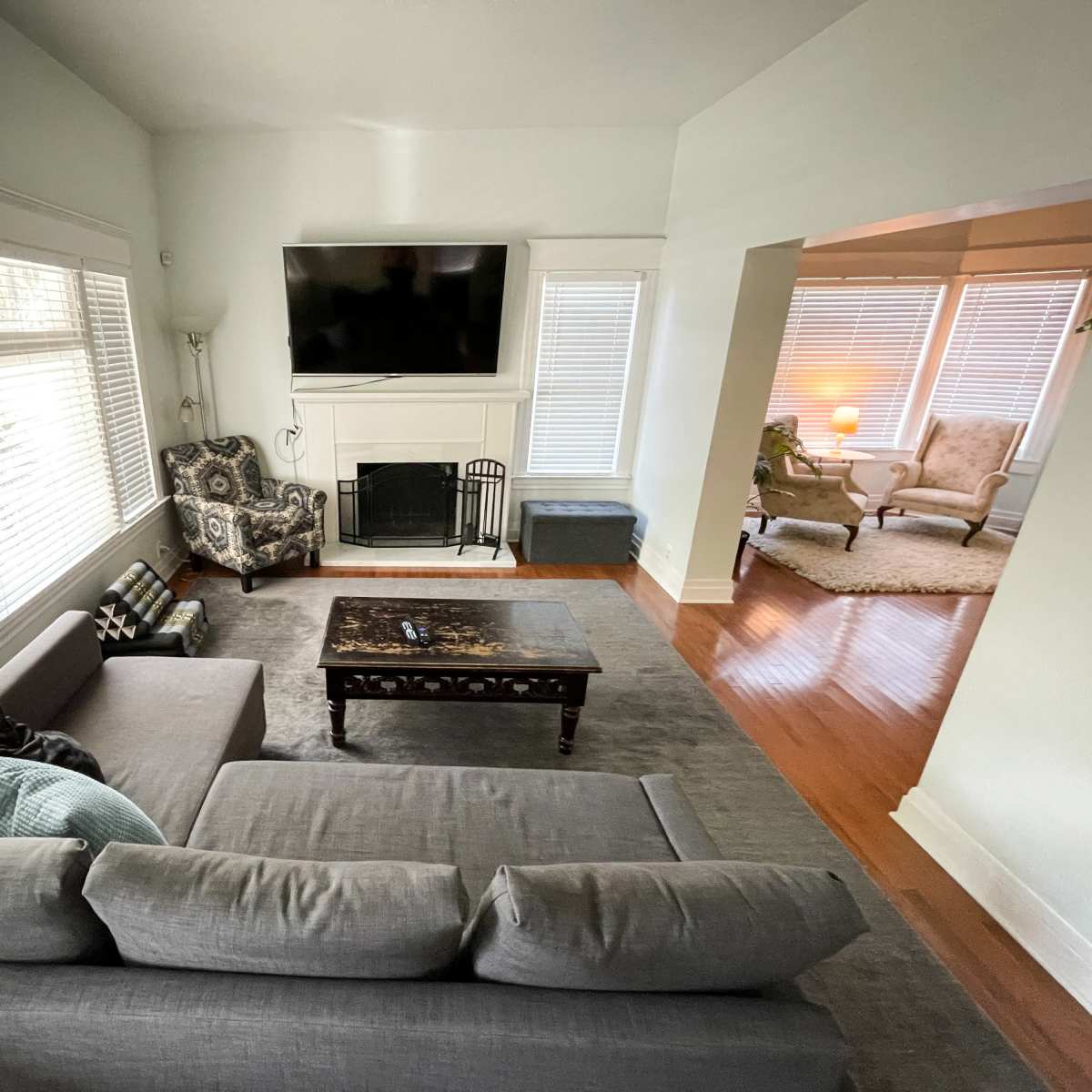 A living room with a gray sectional sofa facing a coffee table and a wall-mounted television, adjacent to a sunlit area featuring additional seating.